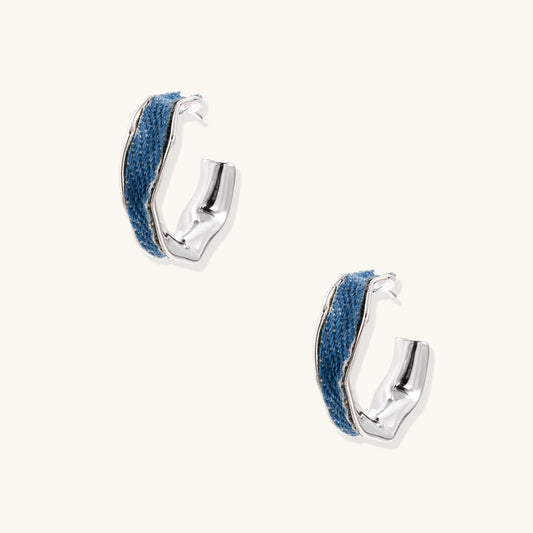 Silver huggie hoops with blue denim fabric inlay strip and polished white gold plated finish on cream background.